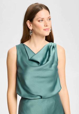 Femme aux longs cheveux bruns portant un haut sans manches en satin bleu sarcelle et des boucles d'oreilles anneaux argentés, regardant légèrement vers la droite.