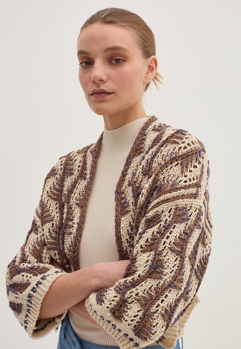 Cardigan lavorato a maglia con base crema, caratterizzato da un motivo a zigzag in marrone e blu. Maniche ampie e bordo decorativo ai polsini.