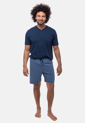 Granatowy T-shirt w serek z kieszonką na piersi, zestawiony z niebieskimi szortami w kratę na sznurku. Materiał bawełniany, luźny krój, boso.