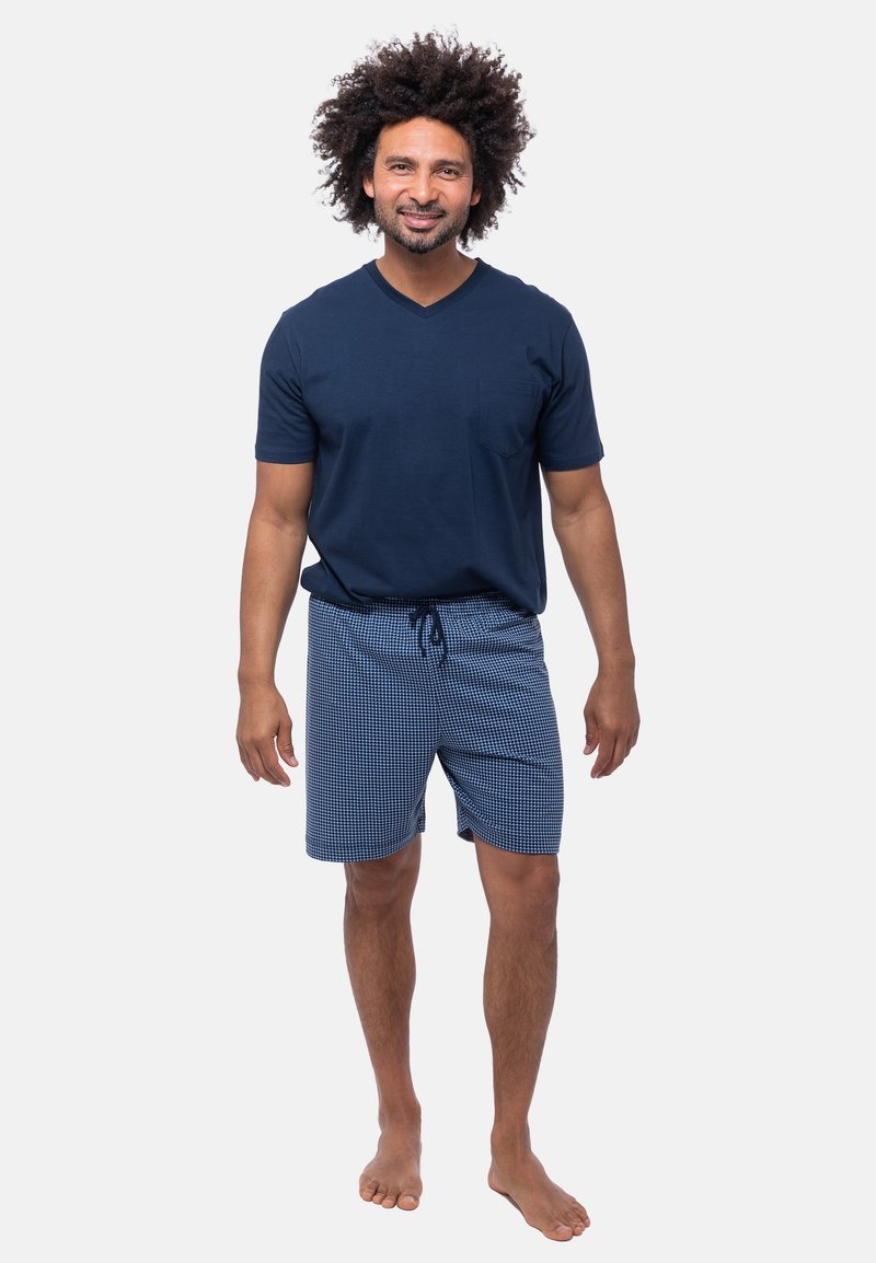 Marineblauwe V-hals T-shirt met een borstzak, gecombineerd met blauw geruite shorts met trekkoord. Katoenen stof, relaxed fit, en blote voeten.