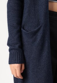 Cardigan largo azul marino, tejido de punto con una textura suave, puños acanalados y bolsillos laterales. El diseño presenta un corte holgado.