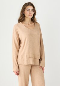 Maglione beige a maglia con maniche lunghe, scollatura a V e dettaglio della tasca frontale. Texture morbida e vestibilità rilassata. Indossato con pantaloni abbinati.