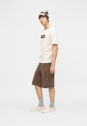 Νεαρός άνδρας που στέκεται, φοράει ένα λευκό μπλουζάκι με γραφικά, καφέ cargo σορτς, γκρι κάλτσες, παπούτσια χωρίς κορδόνια και ένα καφέ πλεκτό σκουφάκι με σχέδιο.