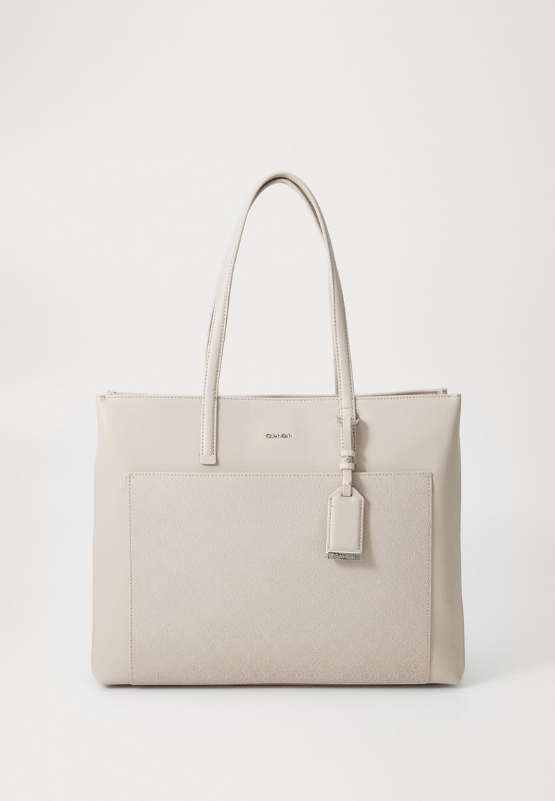 Sac fourre-tout beige Calvin Klein avec doubles poignées, poche texturée à l'avant et une étiquette en cuir suspendue sur un fond blanc uni.