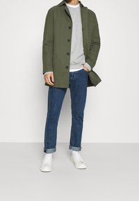 Olivgrön kappa med krage och fem knappar, tillsammans med en grå sweatshirt och blå jeans. Vita sneakers fulländar outfiten.