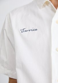 Short-sleeved, white cotton shirt with a relaxed fit, featuring blue embroidered text "Service" on the left side of the chest.