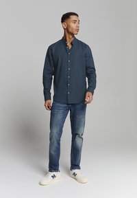 Marineblauwe button-up shirt met een bandkraag, gecombineerd met blauwe jeans en witte sneakers met blauwe accenten. Gladde stof textuur.