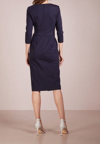 Mörkblå figurnära klänning med trekvartslånga ärmar, knälängd, subtila sömmar och detaljerad dragkedja i ryggen. I kombination med vita högklackade skor.