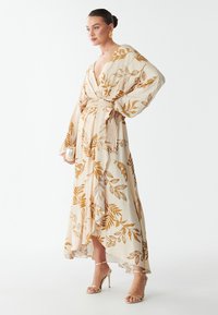 Lange wikkeljurk in crème met gouden bladenpatronen, met ballonmouwen en een riem op de taille. Gekleed met sleehakken.
