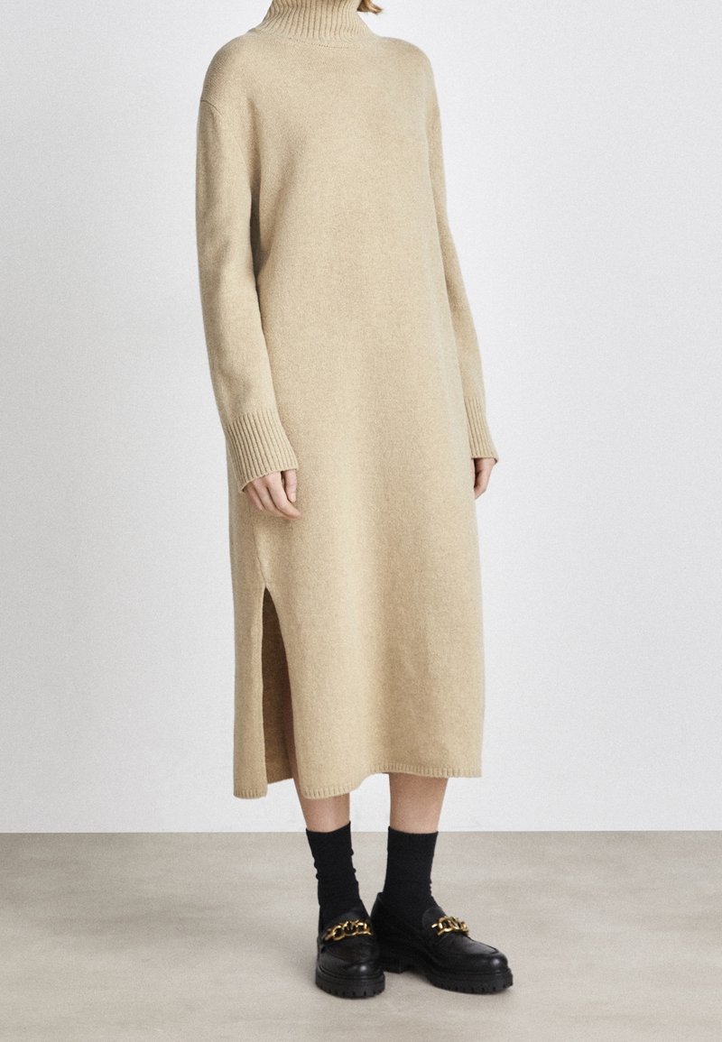 Robe longue en maille beige à col roulé avec fentes latérales, portée avec des chaussettes noires et des mocassins noirs ornés d’un détail chaîne dorée.