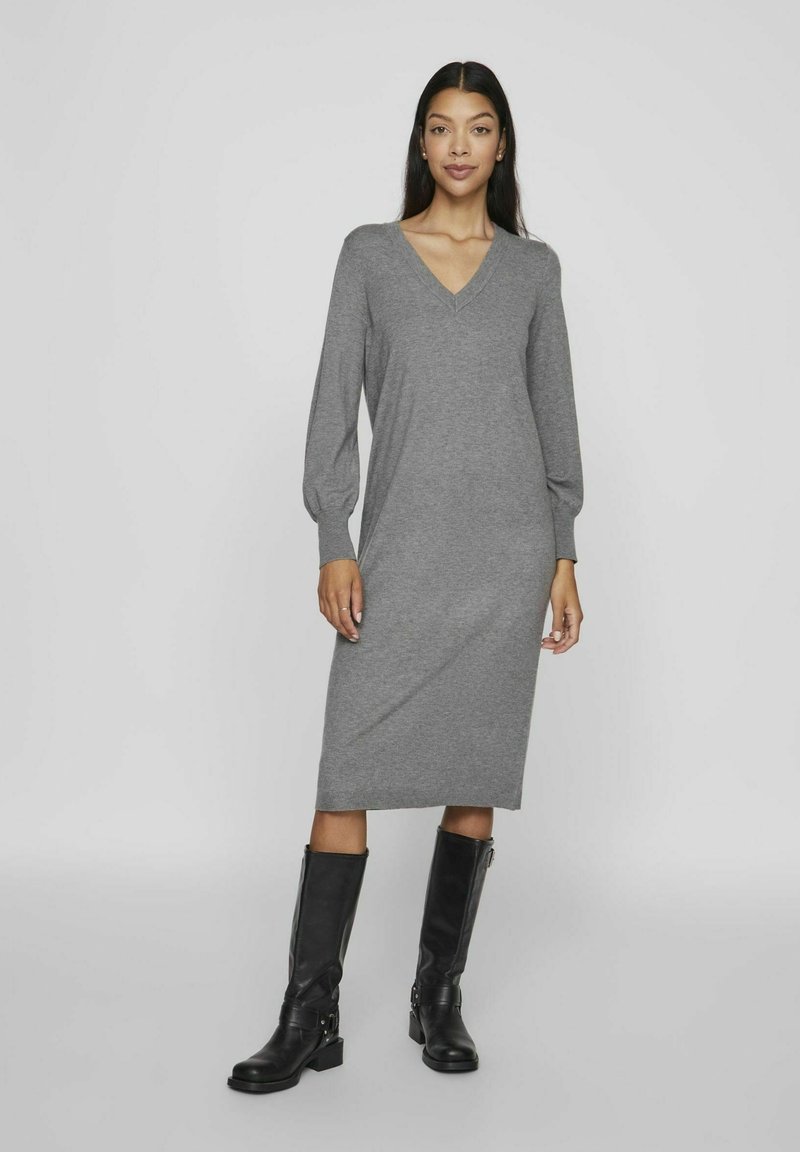 Abito grigio a maglia con scollatura a V, maniche lunghe a sbuffo e silhouette dritta, abbinato a stivali in pelle neri al ginocchio.