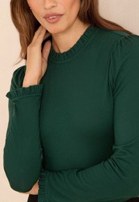 Kobieta ubrana w ciemnozielony top z długim rękawem, z falbankowym kołnierzem i mankietami, delikatnie dotykająca szyi jedną ręką.