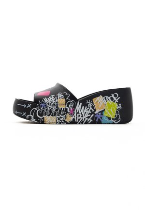 Pieds chaussés de sandales à plateforme noires avec des motifs graffiti colorés, associés à un pantalon noir retroussé sur un fond blanc.