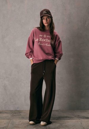 Bordeaux sweatshirt met witte tekst en elastische zoom, gecombineerd met donkere, losse broek en beige schoenen, tegen een grijze achtergrond.