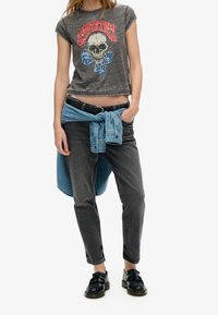 Camiseta gris cortada con un gráfico de calavera, combinada con jeans negros ajustados y una camisa de mezclilla clara atada en la cintura. Zapatos negros complementan el conjunto.