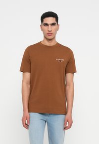 Καφέ βαμβακερό μπλουζάκι με κοντά μανίκια και λαιμόκοψη. Φέρει ένα μικρό άσπρο λογότυπο στην αριστερή πλευρά του στήθους. Κανονική γραμμή, λεία υφή.