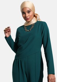 Blusa verde de manga larga con un diseño drapeado y detalles plisados, combinada con un hijab de satén dorado claro y un collar de cadena metálica.
