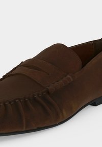 Καφέ μοκασίνι από σουέτ με ραφές moc toe και χαμηλή μαύρη σόλα, εμφανιζόμενο σε κοντινή γωνιακή πλευρική όψη.