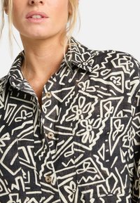 Camisa de patrón en blanco y negro con diseños abstractos, tapeta de botones y cuello. La tela parece ligera con un acabado texturizado.