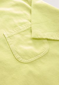 Tessuto di cotone verde lime con superficie testurizzata; presenta una tasca sul petto e una manica piegata, con bordi cuciti e finitura morbida.