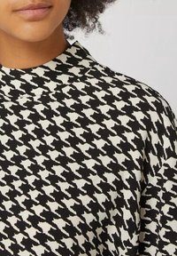 Sort-hvidt houndstooth-mønsterstof med høj krave, der har en tekstureret vævning og abstrakte former i hele designet.
