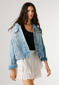 Młoda kobieta o długich ciemnych włosach nosi jasnoniebieską kurtkę dżinsową na czarnym topie oraz warstwową białą spódnicę z falbanami.