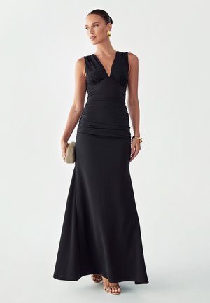 Femme portant une robe longue noire sans manches, tenant une pochette beige, accessoirisée avec des boucles d'oreilles, un bracelet et des sandales dorés.