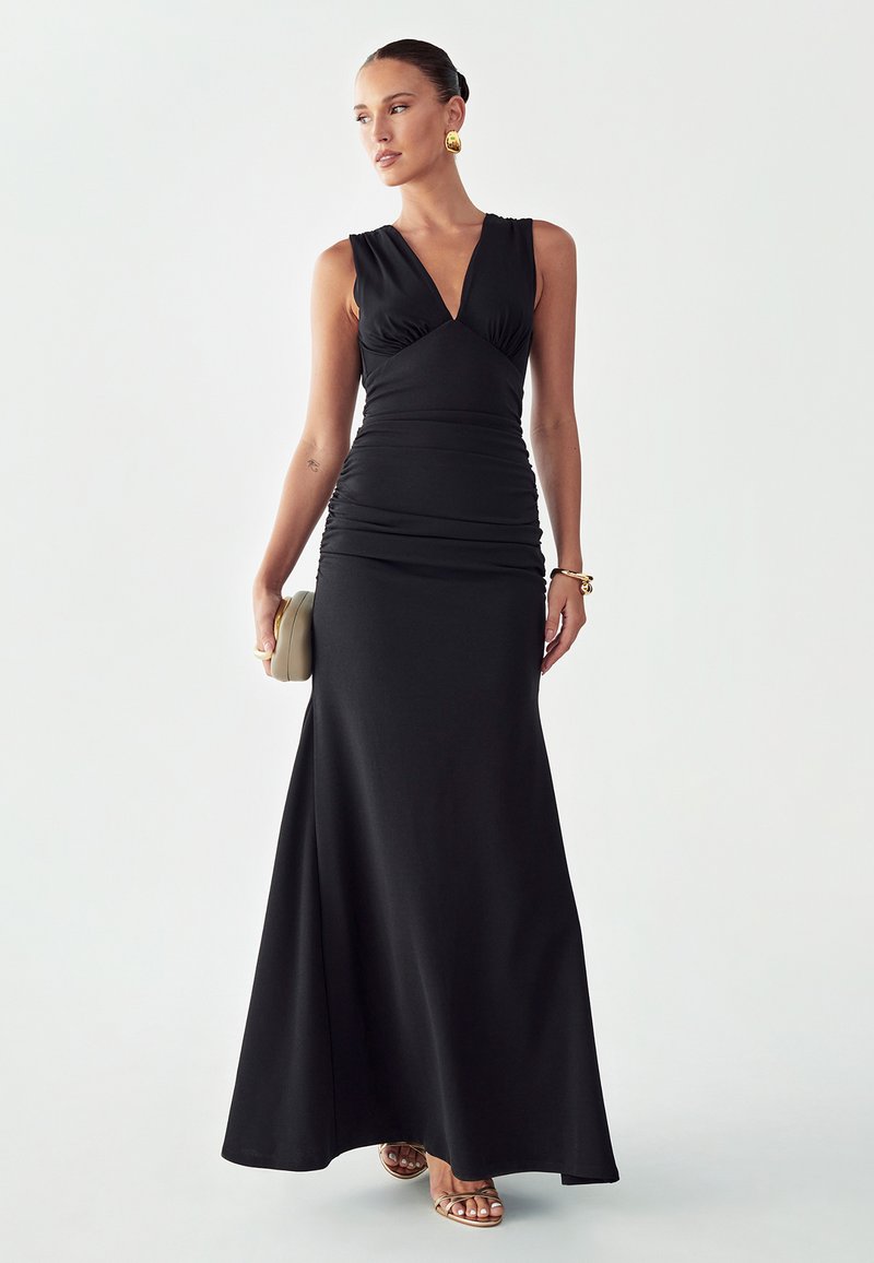 Frau in einem ärmellosen, bodenlangen schwarzen Kleid, die eine beige Clutch hält und mit goldenen Ohrringen, Armband und Sandalen accessoriert ist.