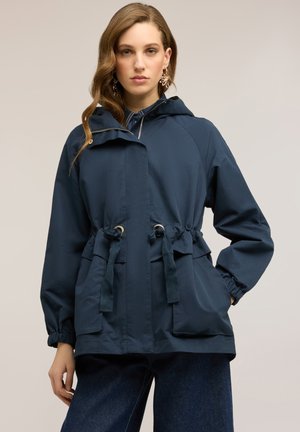 Mujer con chaqueta con capucha azul marino con cintura ajustable y bolsillos frontales, combinada con vaqueros azul oscuro, de pie sobre un fondo liso.