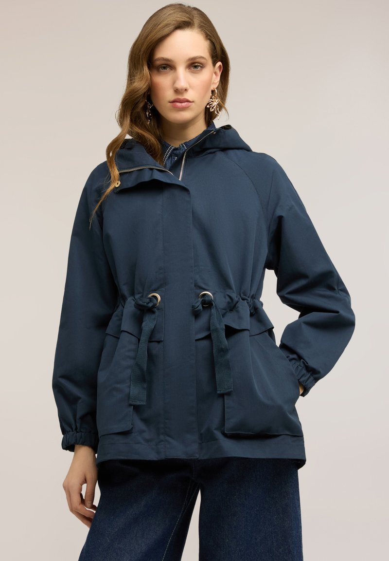 Mujer con chaqueta con capucha azul marino con cintura ajustable y bolsillos frontales, combinada con vaqueros azul oscuro, de pie sobre un fondo liso.