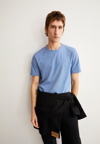 Junger Mann mit kurzen braunen Haaren trägt ein hellblau gestreiftes T-Shirt und schwarze Hosen, mit einer schwarzen Jacke um die Taille gebunden, stehend vor einer weißen Wand.