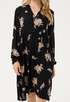 Zwarte lange mouwen V-hals jurk met beige bloemenpatronen, geplooide taille en elastische manchetten gedragen door een persoon met blond haar.