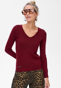 Bordowy sweter w serek o prążkowanej fakturze. Połączony z spodniami w panterkę. Widoczna brązowa metka marki w dolnym lewym rogu. Na nosie duże okulary przeciwsłoneczne.