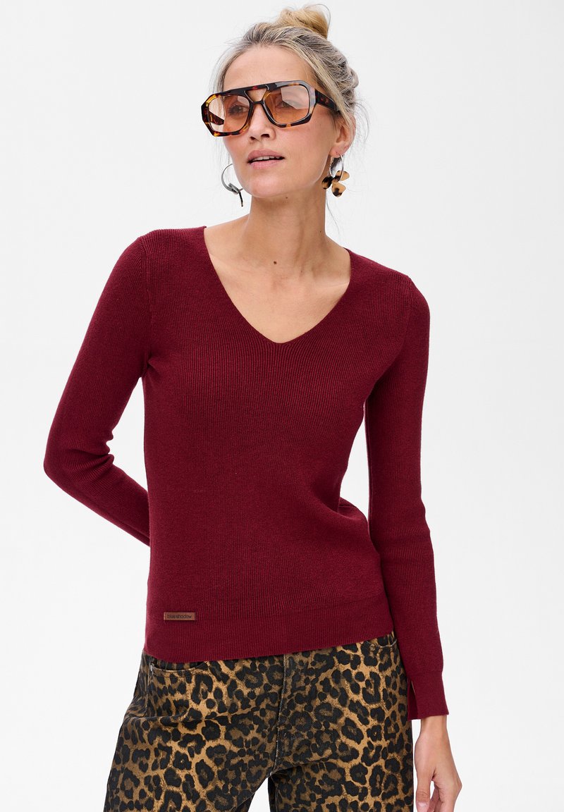 Bordowy sweter w serek o prążkowanej fakturze. Połączony z spodniami w panterkę. Widoczna brązowa metka marki w dolnym lewym rogu. Na nosie duże okulary przeciwsłoneczne.