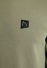 Gros plan sur un tissu en maille gaufrée vert olive avec un petit patch carré noir affichant deux lignes blanches inclinées et superposées.