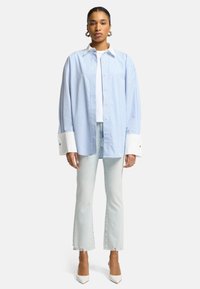 Camicia oversize a righe blu e bianche con colletto bianco, dettagli con bottoni e polsini larghi, abbinata a jeans azzurri e tacchi bianchi.
