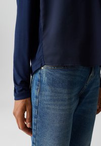 Marineblaues Langarmoberteil mit einer glatten, leichten Textur, kombiniert mit mittelhohen blauen Jeans aus Denim, die eine dezente Waschung und Kontrastnähte aufweisen.