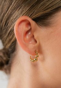 Goldene Creolen mit einem geschwungenen Design und unterschiedlich großen sphärischen Akzenten, in Nahaufnahme zusammen mit einem strukturierten Ohr präsentiert.