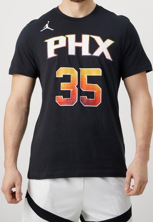 Mężczyzna w czarnej koszulce koszykarskiej PHX z numerem 35 w gradientowym pomarańczowym kolorze, białe spodenki z czarnym ukośnym paskiem, stoi na jasnym tle.