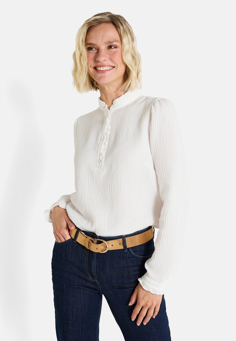 Blusa blanca con volantes, de manga larga y cierre de botones, combinada con jeans de mezclilla oscuros y un cinturón marrón con hebilla redonda.