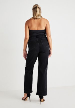 Woman with blonde hair in a half-up style wearing a black strapless top, high-waisted black jeans, and black high-heeled sandals, standing back.