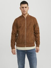 Chaqueta bomber de ante marrón con puños y cuello acanalados, con cremalleras plateadas y bolsillos laterales. Llevada sobre un suéter de color crema claro.