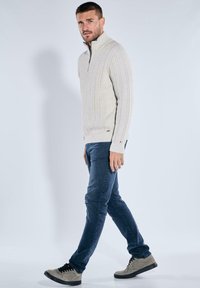 Hellgrauer, gerippter Strick-Zip-Pullover mit hohem Kragen, kombiniert mit dunkelblauen Jeans und beigen Sneakern, was ein lässiges Outfit ergibt.