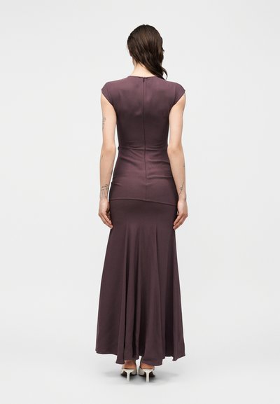 Ženska z dolgimi temnimi lasmi, oblečena v oprijeto vijolično dolgo obleko in bele pete, stoji z obrnjenimi hrbtom proti kameri na enobarvnem ozadju.