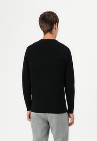 Pull noir en maille avec des manches longues et un col rond. Le tissu semble doux et texturé, associé à un pantalon gris à rayures.