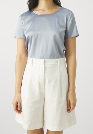 Vrouw draagt een lichtblauw topje met korte mouwen en witte knielange shorts met hoge taille, staand tegen een effen achtergrond.
