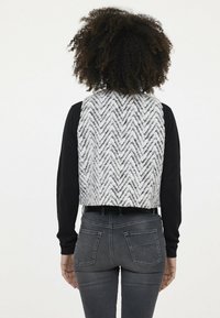 Grauer und weißer Strickwesten im Fischgrätenmuster, gekürzte Ausführung aus dickerem Stoff, getragen über langen schwarzen Ärmeln und hoch taillierten grauen Jeans.