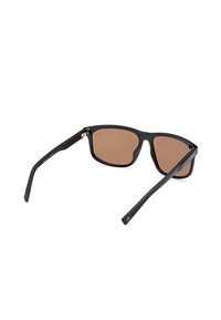 Schwarze Sonnenbrille mit braun getönten Gläsern. Geometrisches Design, schlanke Bügel mit strukturierten Griffen. Metallelemente am Scharnier.