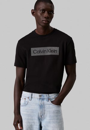 Camiseta negra de manga corta con un parche texturizado del logo de Calvin Klein. Combinada con jeans de mezclilla azul claro. El modelo se encuentra de pie contra un fondo gris.