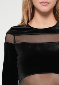 Top preto de mangas compridas com material de veludo, um painel de malha transparente e decote redondo. Acentos com elementos estruturados ao longo dos ombros.